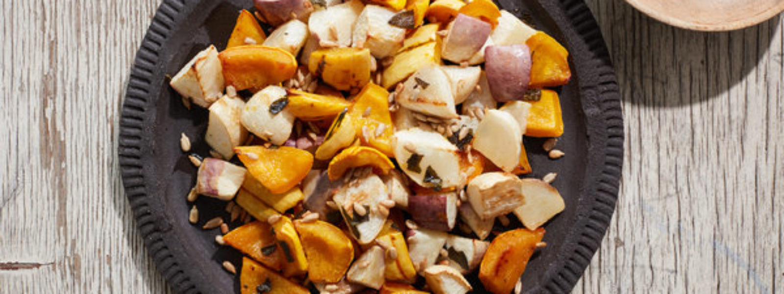 A picture of Roasted Turnips and Winter Squash With Agave Glaze from directly above. The orange and pale root vegetables sit on a slate grey plate with a brown bowl full of a darker brown grain sit in the upper right hand corner of the image.
