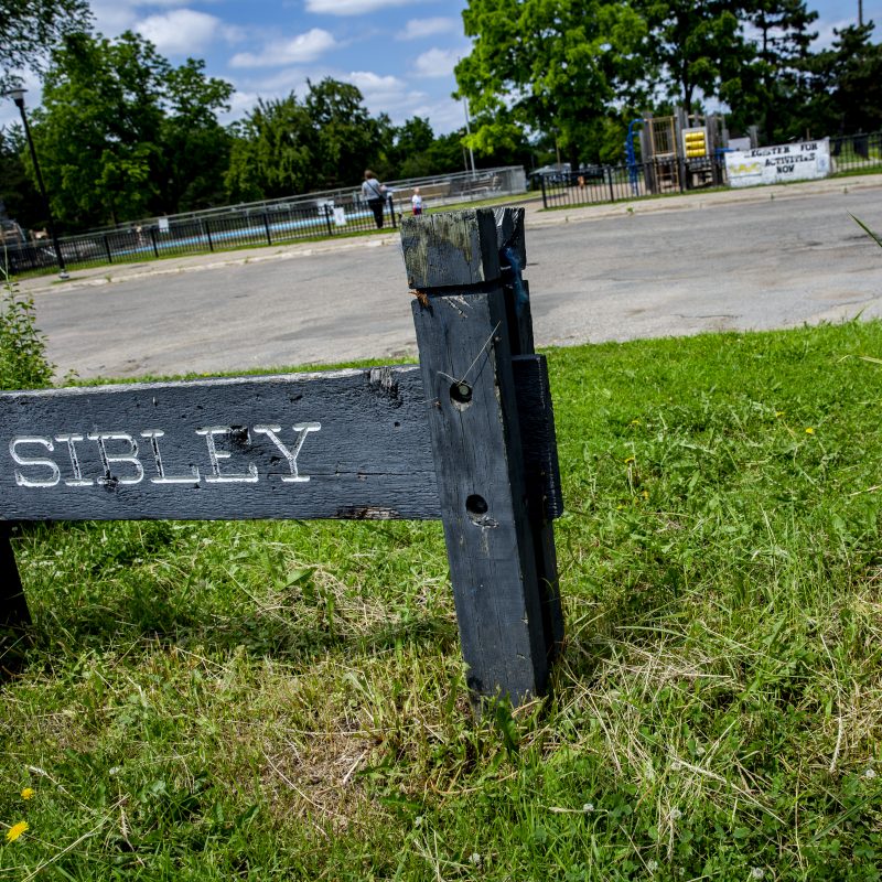 Wooden sign that says "Sibley" standing in the grass in front of the Sibley Park Parking Lot